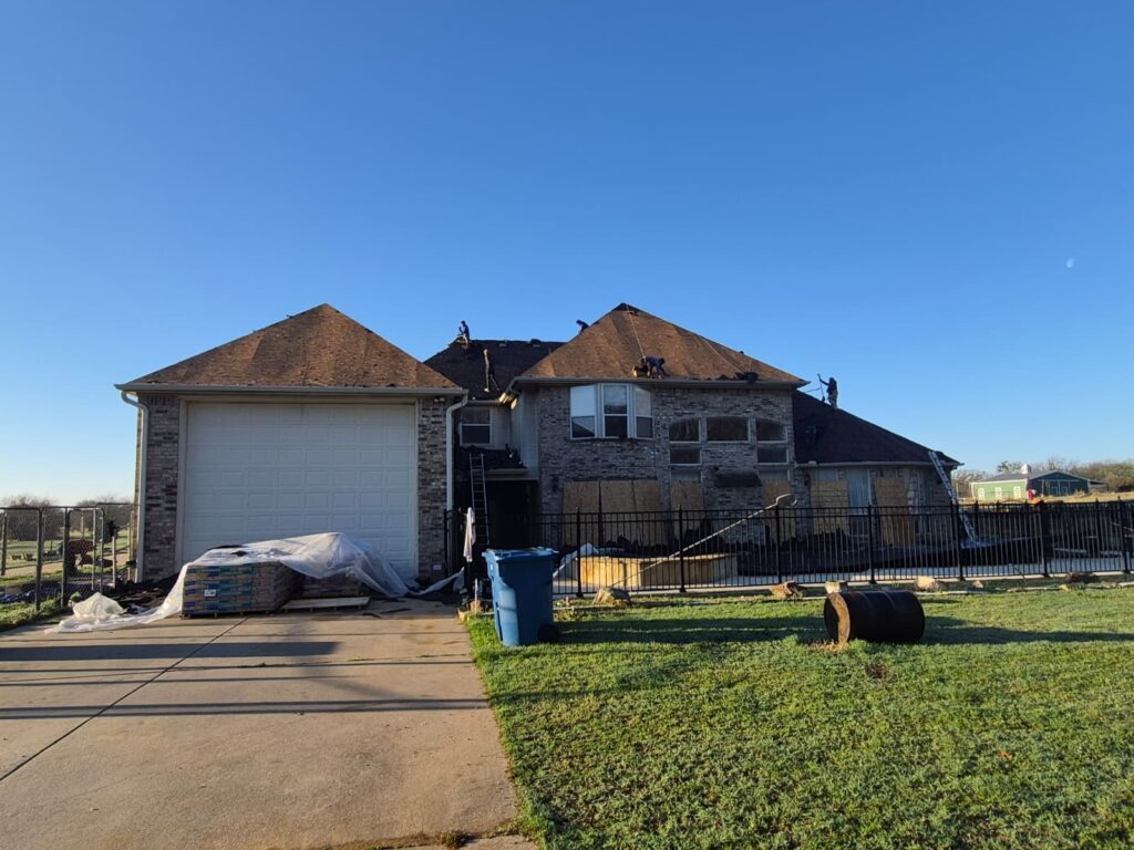 A crew is actively working on the roof of a large residential house under a clear blue sky. Bundles of new roofing material are visible on the driveway, and some windows on the house are boarded up.