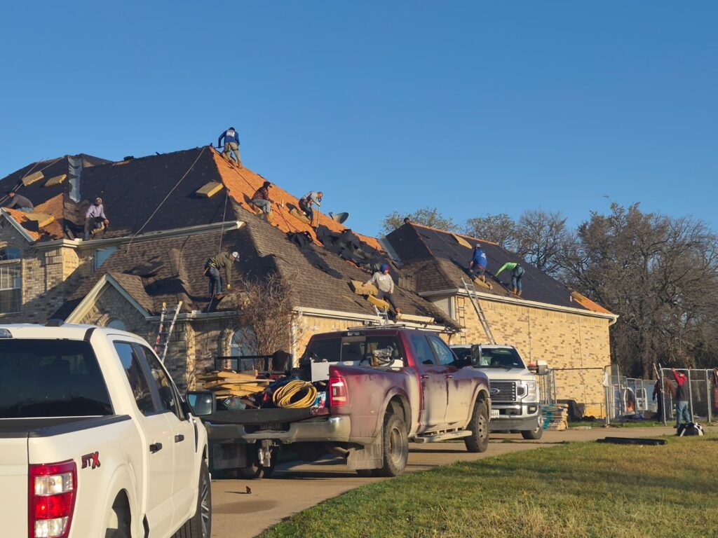 A crew of roofers is actively working on a residential brick home, removing old shingles and installing new roofing materials, with several work trucks parked nearby.