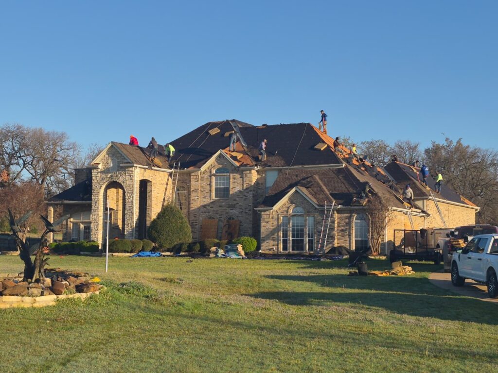 A large, two-story brick and stone house with a crew of roofers actively working on replacing the asphalt shingle roof. The roof is partially stripped down to the underlayment and partially re-shingled, with ladders and debris visible around the property.