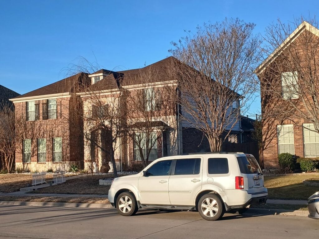 A large brick house with a brown asphalt shingle roof is seen from the street, with a white SUV parked in front and bare trees. The sky is clear blue.