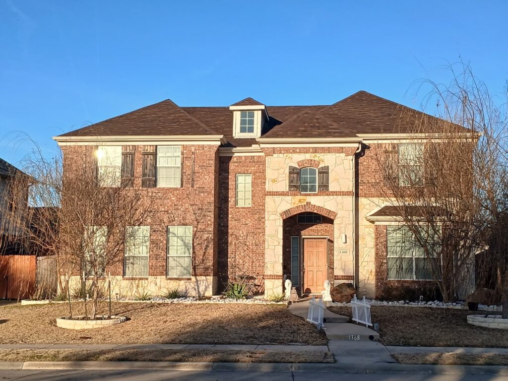 A two-story residential house with a brown asphalt shingle roof, brick and stone facade, and a clear blue sky overhead. The house is viewed from the street, showing the front yard and entrance.