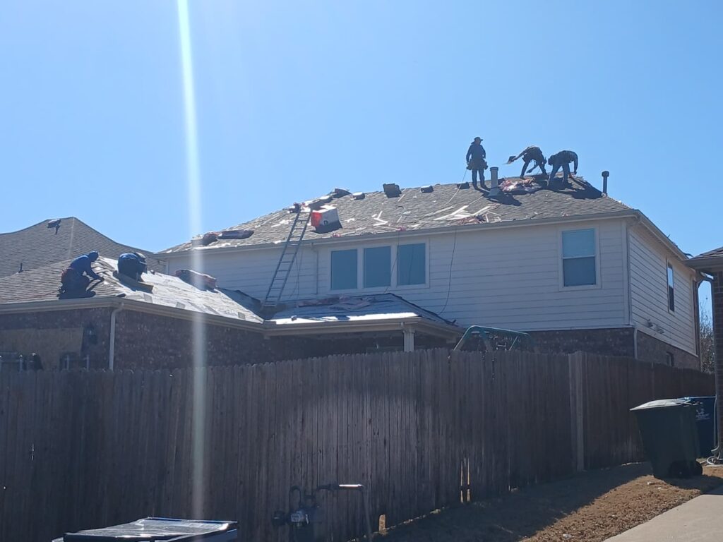 Several roofers are actively working on a residential roof, which is in the process of being replaced with old shingles removed and underlayment visible. A ladder leans against the house, which features light siding and brick, with a wooden fence in the foreground.