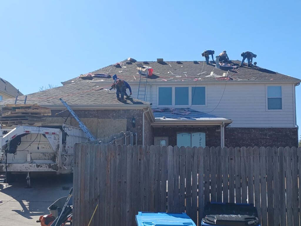 Several roofers are actively working on a residential roof, which appears to be in the process of being replaced. A dump trailer is parked in the driveway below, and a wooden fence partially obscures the lower portion of the house.