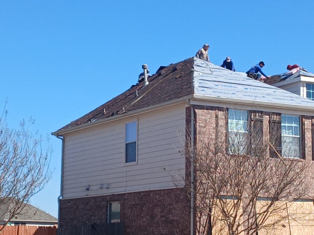 Several roofers are actively working on a residential roof, with some sections showing old asphalt shingles being removed and other sections covered with new underlayment.