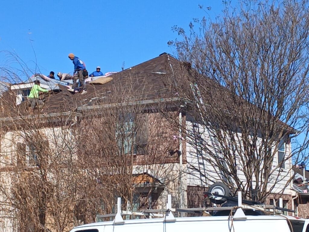 Several roofers are actively working on the brown asphalt shingle roof of a residential house, partially obscured by bare trees in the foreground. A white work truck with a ladder rack is visible at the bottom of the frame.