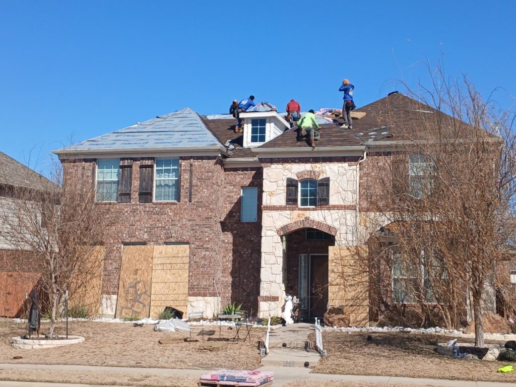 A crew of roofers is actively working on the roof of a large residential house, with some sections covered in underlayment and others with new shingles. The house features brick and stone siding, and several ground-level windows are boarded up.