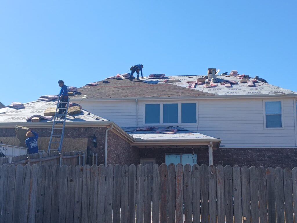 Roofers are actively working on replacing the asphalt shingle roof of a residential house, with bundles of new shingles and underlayment visible on the roof deck.