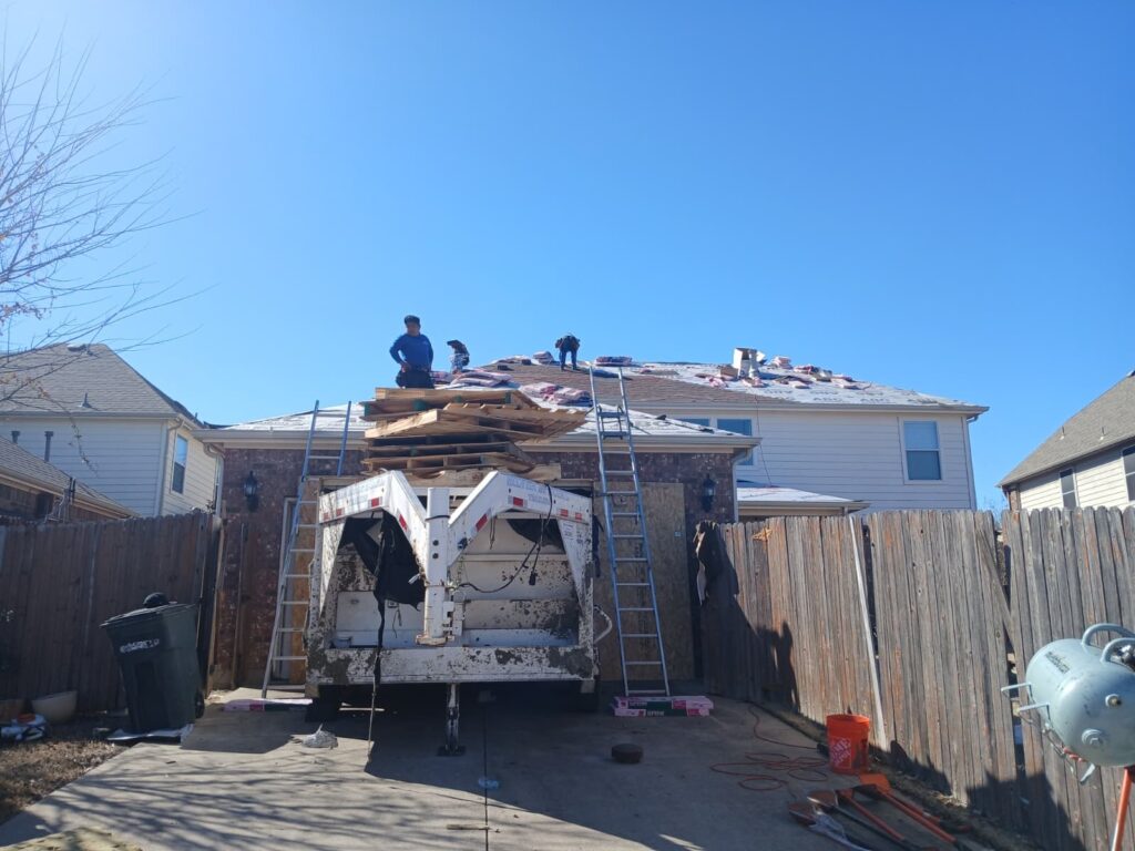Several roofers are actively working on a residential roof, which is partially stripped with new bundles of shingles visible. A large trailer and ladders are positioned in the driveway, with a clear blue sky overhead.