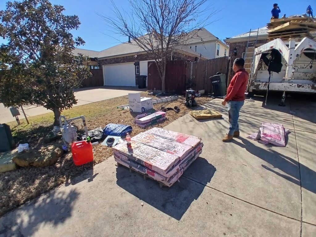 The image shows a residential driveway with stacks of asphalt shingles on a pallet, a worker standing in the foreground, and other crew members actively working on the roof of the house, next to a dump trailer.