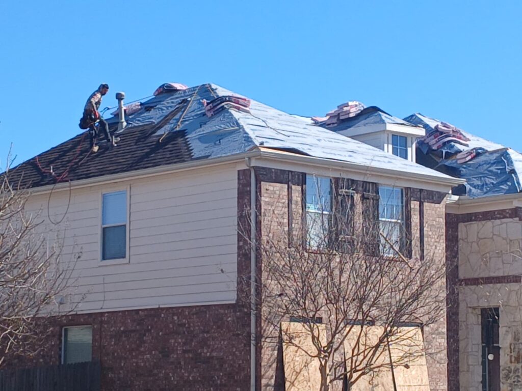 A roofing worker is on a residential roof that is partially stripped, showing underlayment and stacks of new shingles. The house features brick and siding, with some windows boarded up, under a clear blue sky.