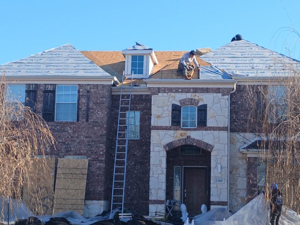 Roofers are actively working on a residential roof, which is partially covered with underlayment and has exposed plywood decking. A ladder is propped against the house, and debris is visible on the ground.