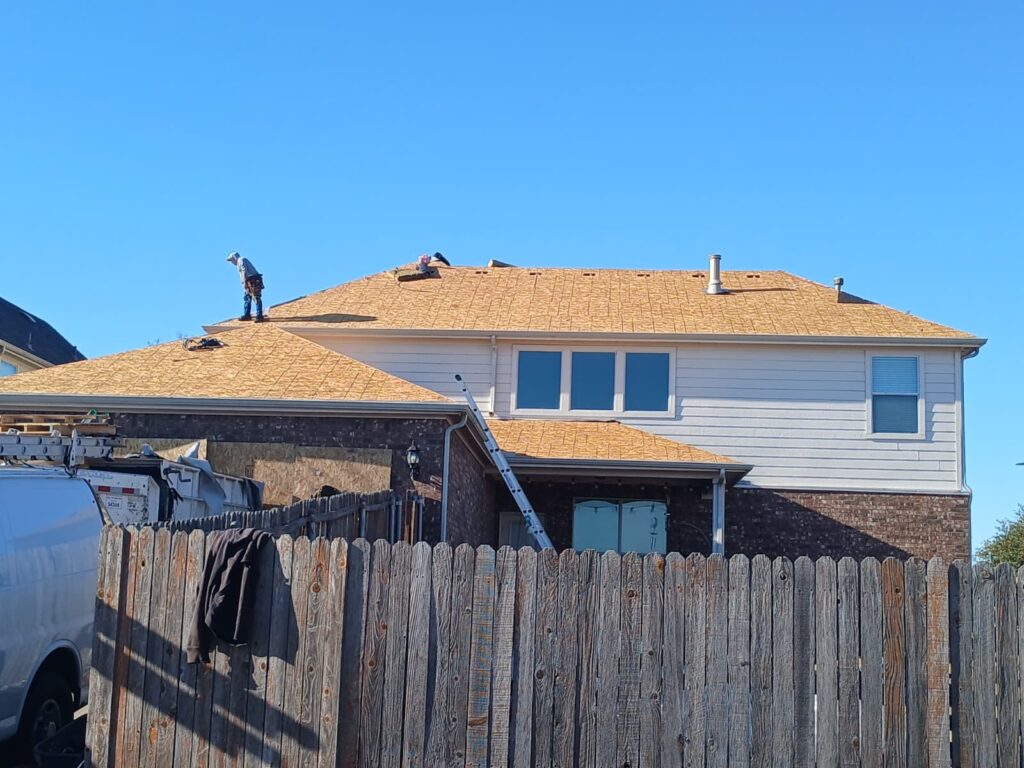 Two roofers are actively working on the roof of a residential house, which currently shows the underlayment and exposed decking. A ladder leans against the house, and a work truck is partially visible behind a wooden fence.