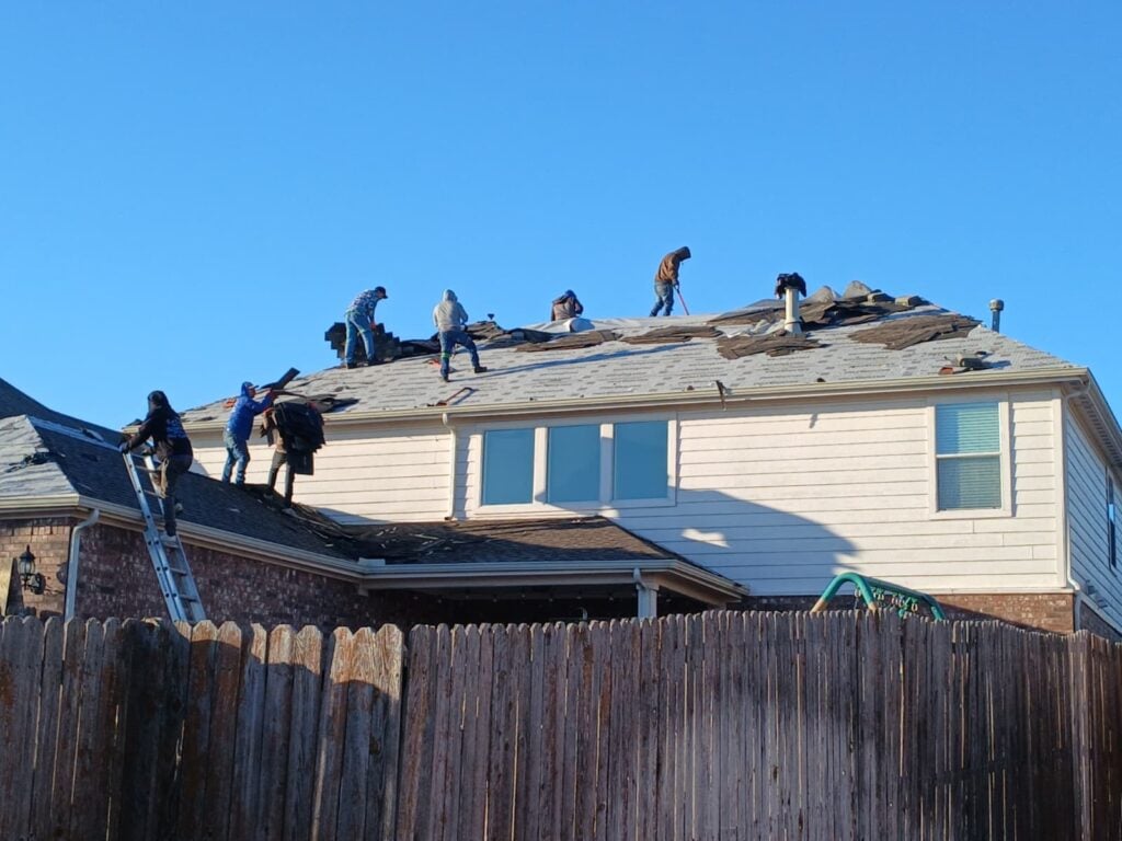 Several roofers are actively working on a residential roof, removing old asphalt shingles under a clear blue sky. One worker is on a ladder, while others are on the roof tearing off material.