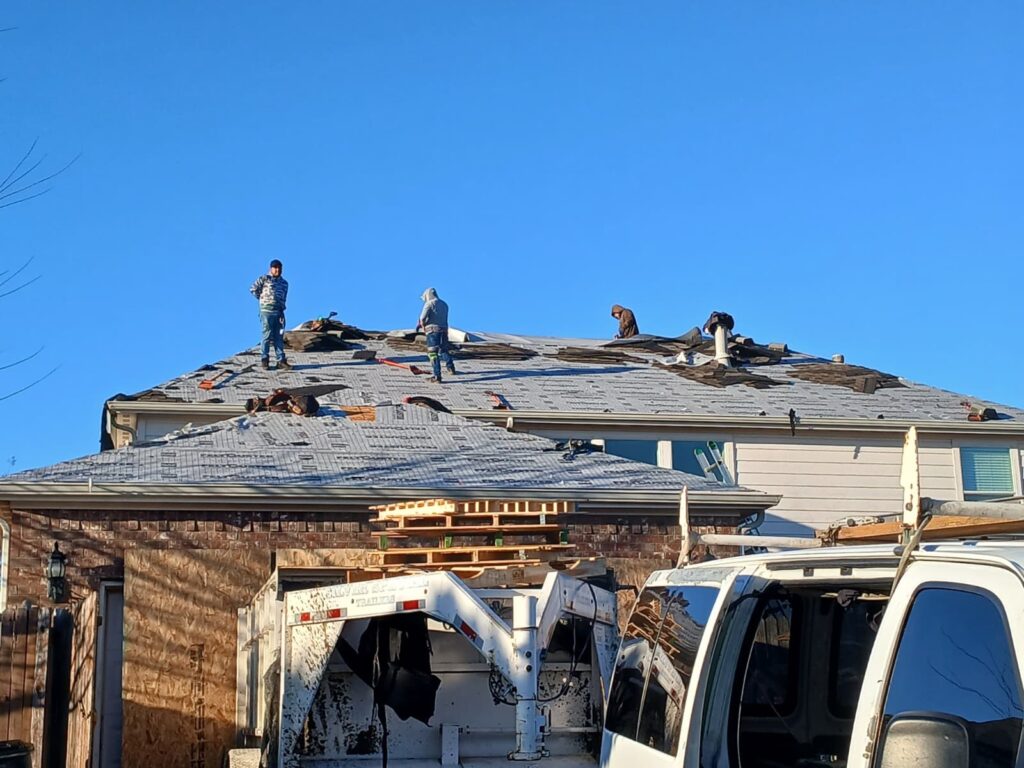 Several roofers are actively stripping old asphalt shingles from a residential roof under a clear blue sky. A dump trailer and part of a work truck are visible in the foreground.
