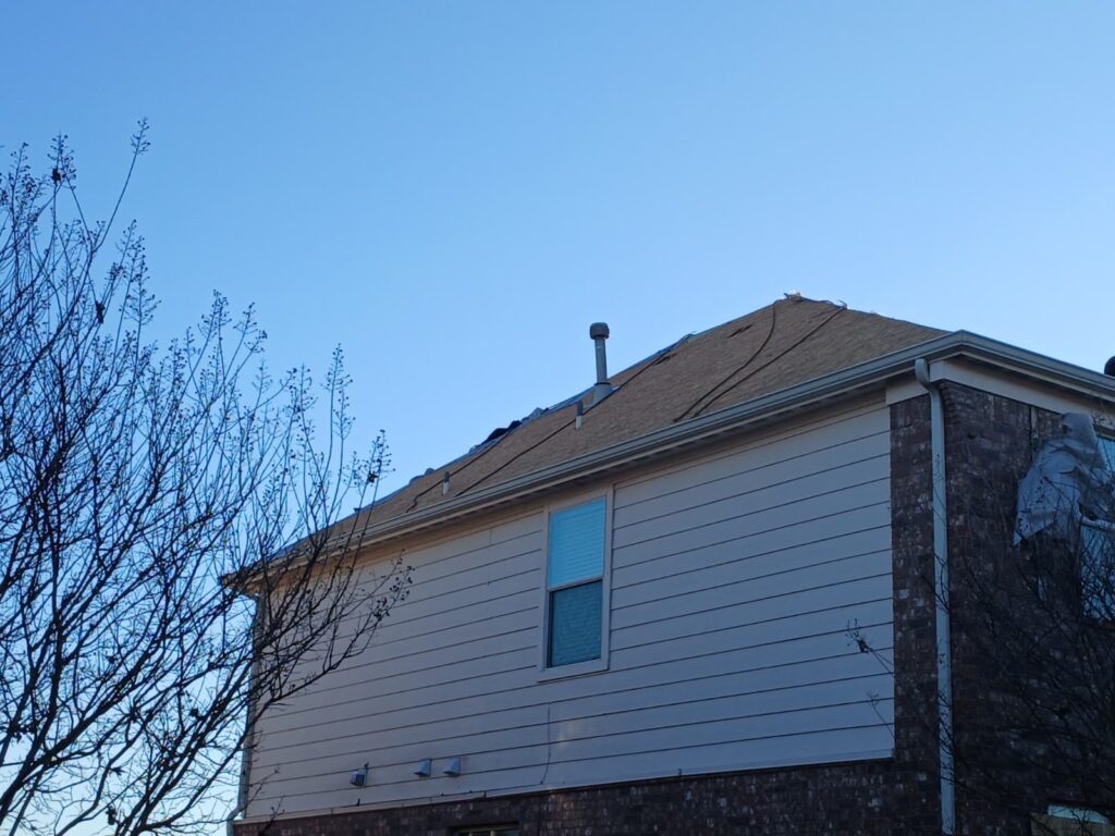 A residential house with a brown asphalt shingle roof partially removed, exposing underlayment. The image shows the side of the house with light-colored siding and brick, under a clear blue sky with bare tree branches in the foreground.