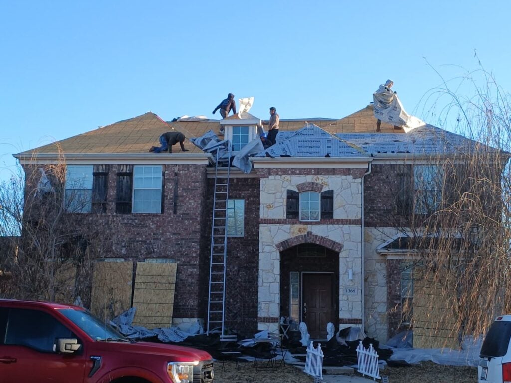 A crew of roofers is actively working on the roof of a large brick and stone residential house, with new underlayment being installed over some remaining old shingles. A ladder is propped against the house, and a red truck is partially visible in the foreground.