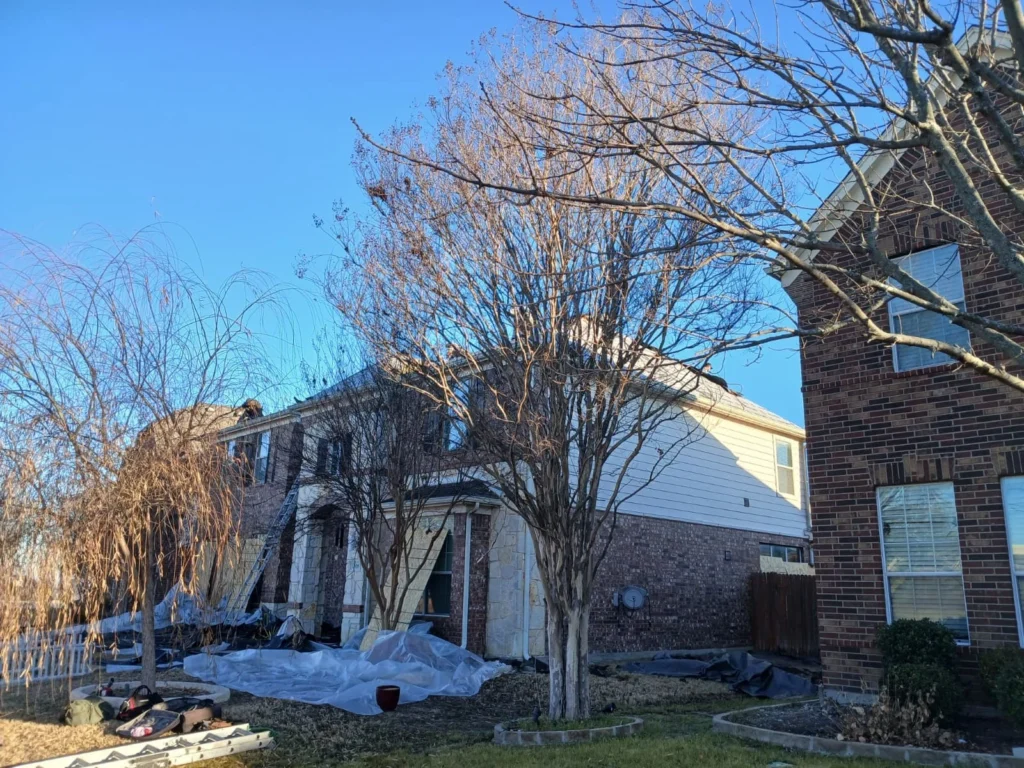 A residential house with roofing work in progress, showing a worker on the roof, a ladder, and protective tarps on the ground. Bare trees are prominent in the foreground and midground.