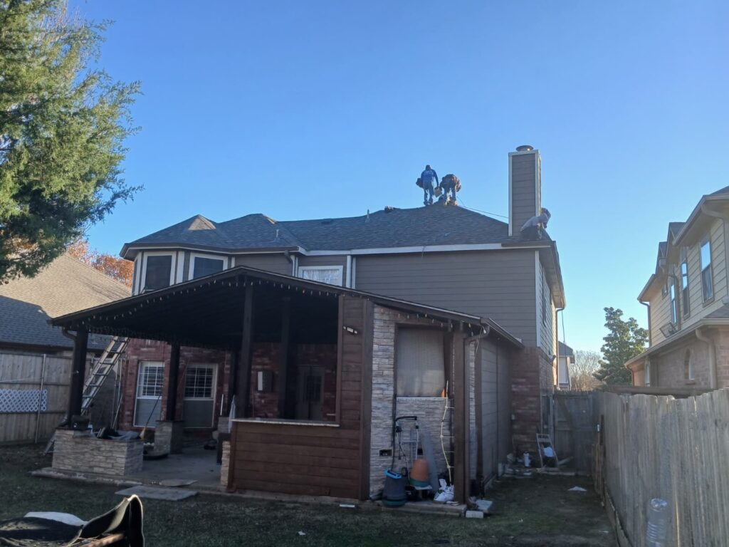 Several roofers are actively working on the asphalt shingle roof of a residential house, with a clear blue sky overhead. The image is taken from the backyard, showing a covered patio area below the main house.