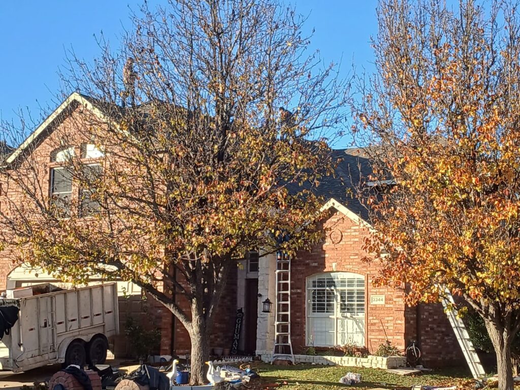 A brick house with a dark asphalt shingle roof is partially obscured by trees with autumn leaves. A worker is visible on the roof, and a dump trailer is parked in the driveway.