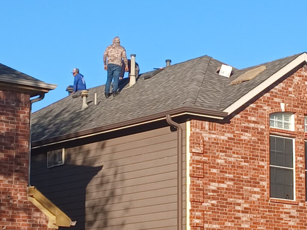 Two roofers are visible on a residential asphalt shingle roof, with one standing and another crouching near a vent. The house features brick and siding, with a clear blue sky in the background.
