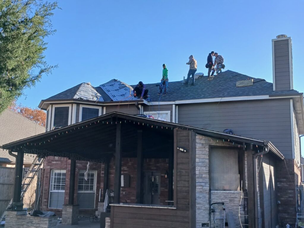 A roofing crew is actively working on a residential roof, with some sections covered in new asphalt shingles and other areas showing the underlayment. The house features a mix of brick, stone, and siding, with Christmas lights strung on the porch.