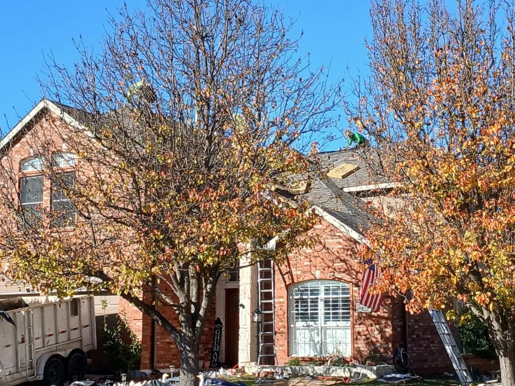 An autumn scene showing a brick house with two roofers working on the asphalt shingle roof, partially obscured by trees with fall foliage. A dump trailer is visible on the left side of the house.