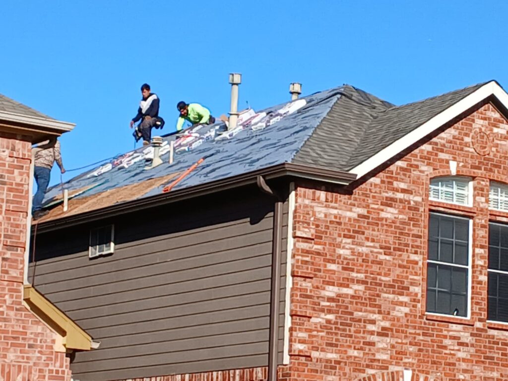Three roofers are actively working on a residential roof, with some underlayment exposed and new asphalt shingles partially installed. The house features brick and siding, under a clear blue sky.
