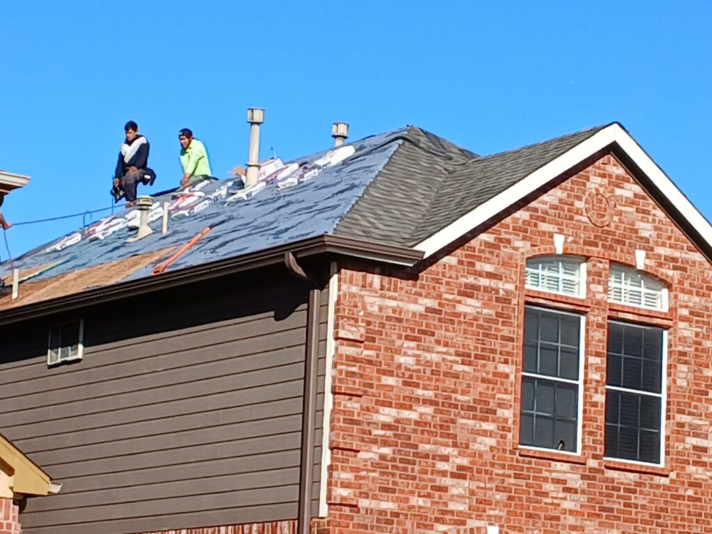 Two roofers are actively working on a residential roof, which is partially stripped with underlayment visible and new shingle bundles ready for installation. The house features a combination of brick and siding.