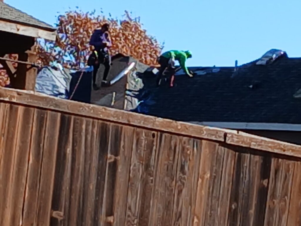 Two roofers are visible on a residential roof, partially obscured by a wooden fence in the foreground. They appear to be actively working on the asphalt shingle roof, with some underlayment or debris visible.