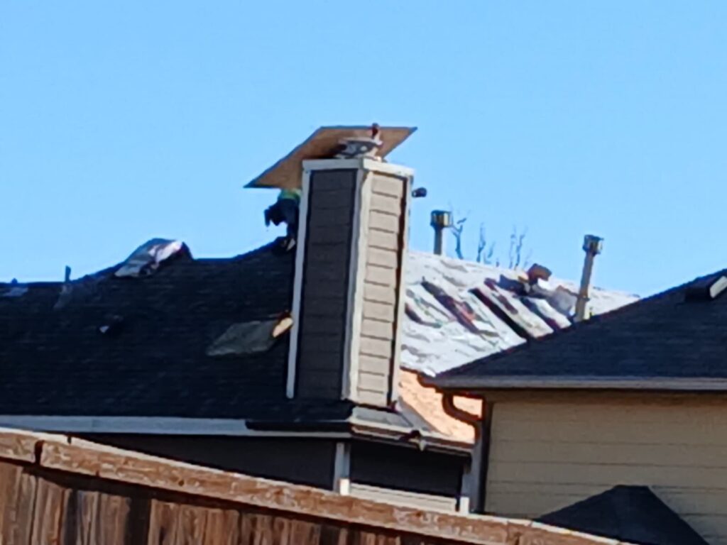 A blurry image showing a worker on a residential roof near a chimney, with parts of the roof covered in underlayment and other parts with new asphalt shingles.