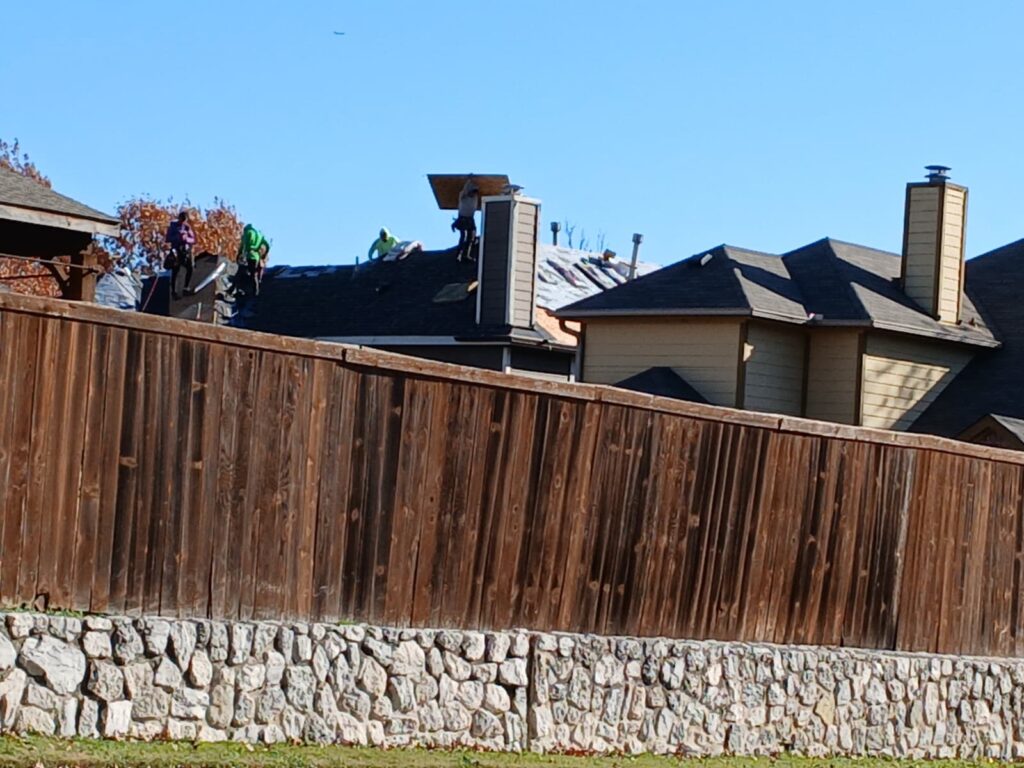 Several roofers are actively working on the roof of a residential house, with some underlayment visible. The lower portion of the image is dominated by a wooden fence and a stone wall.