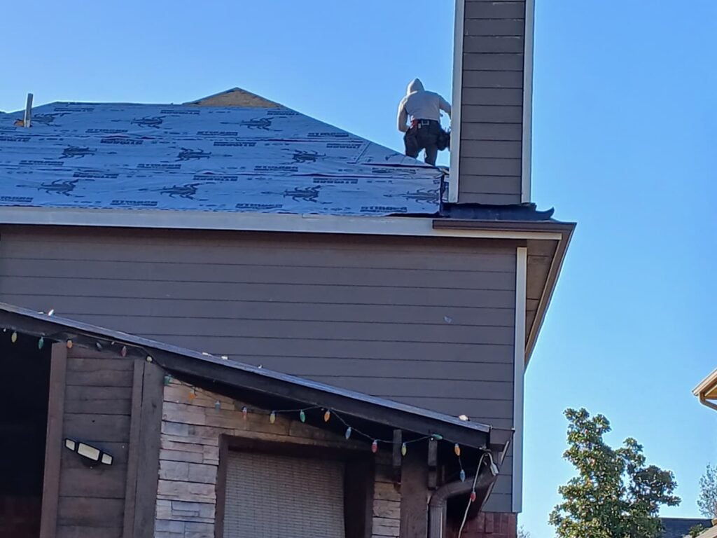 A roofer in a hooded sweatshirt and tool belt is working on a residential roof that is currently covered with blue synthetic underlayment, near a chimney. Part of the house's siding and stone facade is visible below, adorned with colorful string lights.