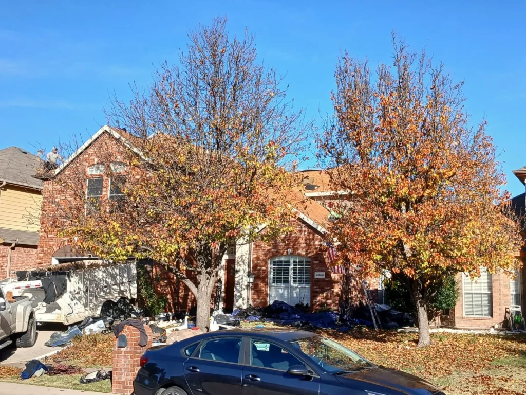 A brick house with a partially removed asphalt shingle roof, showing a worker on the roof and debris on the ground covered by tarps. Two trees with autumn leaves partially obscure the view of the house.