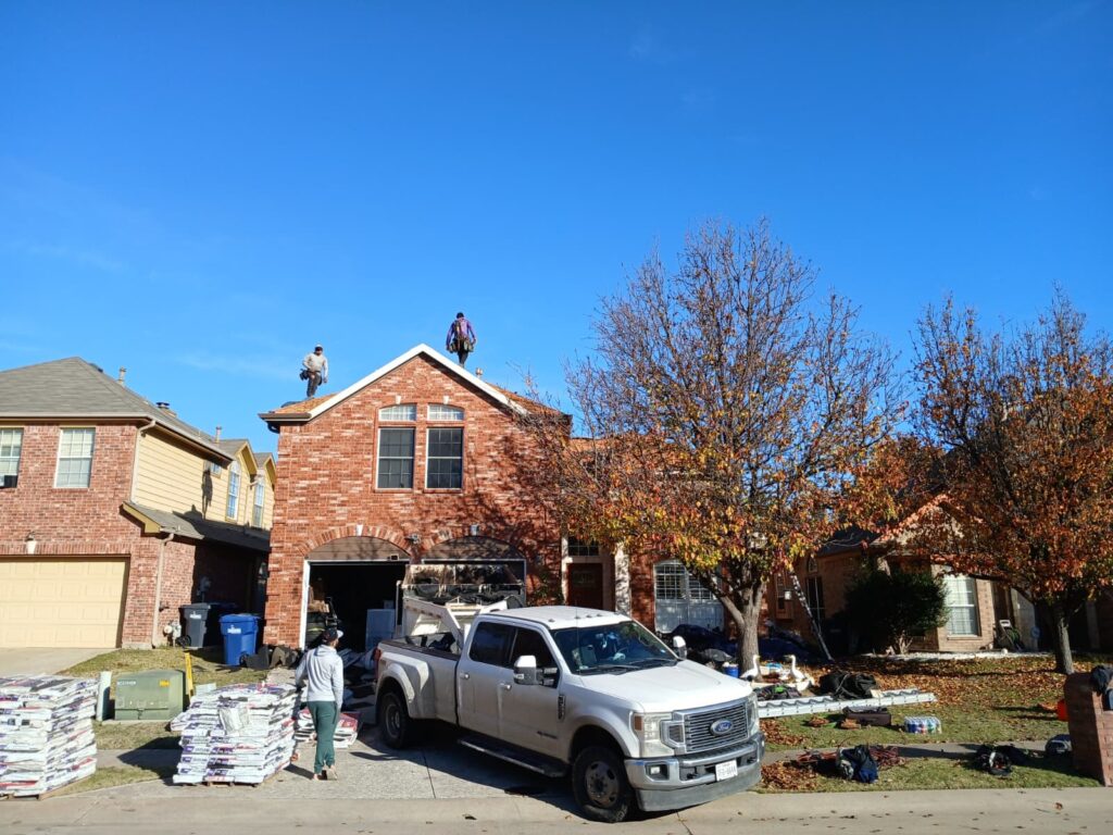 Two roofers are visible on the asphalt shingle roof of a brick residential house under a clear blue sky. A white Ford work truck is parked in the driveway, with pallets of roofing materials stacked nearby.