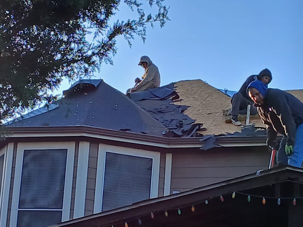 Several roofers are actively working on a residential roof, stripping old asphalt shingles and installing new material on a sunny day.