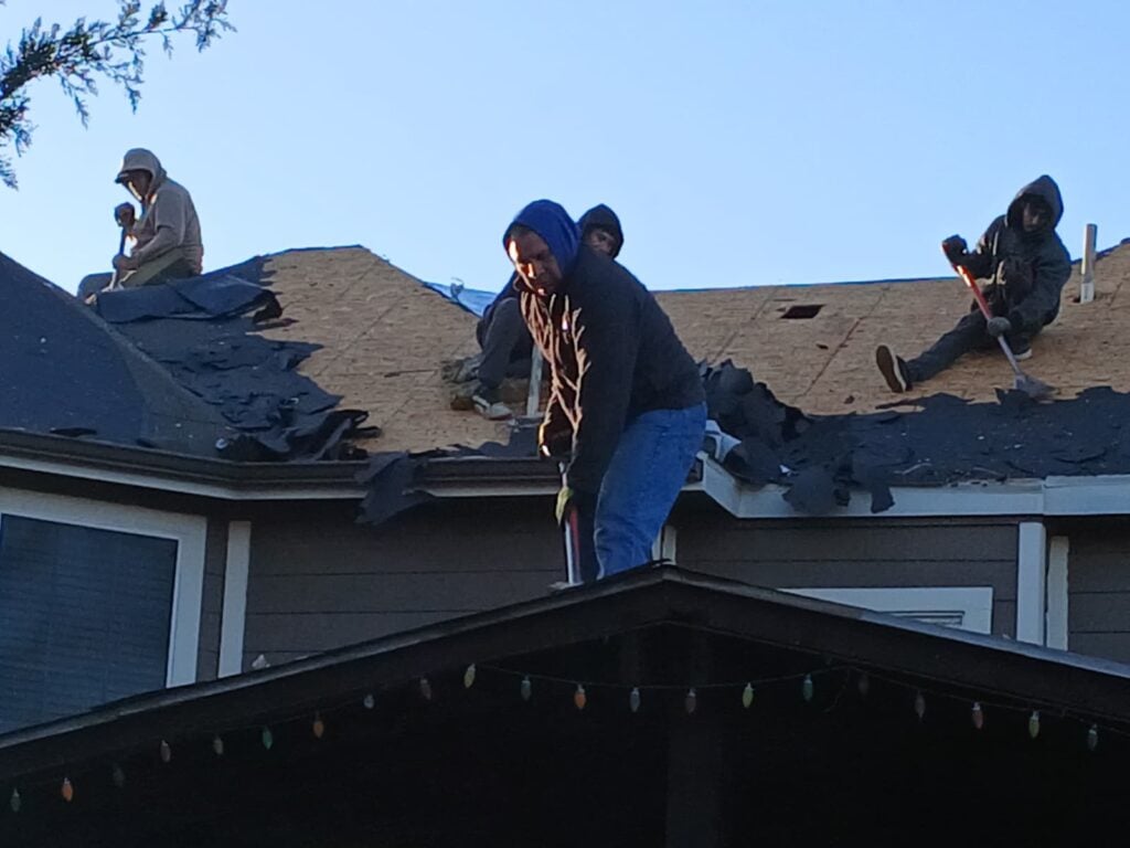 Several roofers are actively removing old asphalt shingles from a residential roof, exposing the underlying roof deck. The workers are wearing hoodies and using tools to tear off the old material.