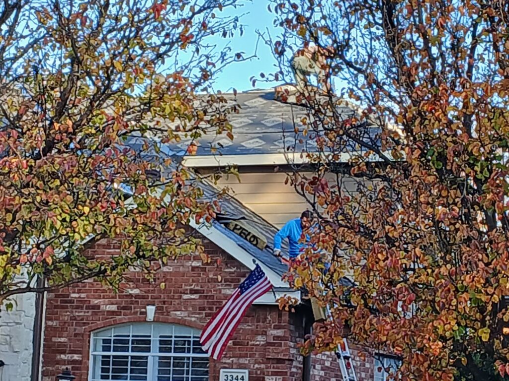 A roofing worker in a blue shirt is visible on a residential roof, which is partially covered with underlayment and new shingles. The scene is framed by autumn trees with colorful leaves, and an American flag hangs from the house below.