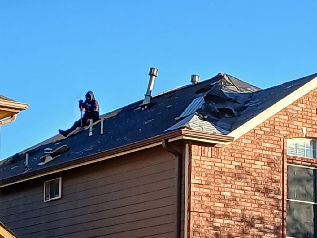 A person wearing a hooded sweatshirt is sitting on a severely damaged asphalt shingle roof, holding a tool, likely in the process of removing old shingles or inspecting damage. The roof shows extensive tearing and missing shingles, exposing the underlayment.
