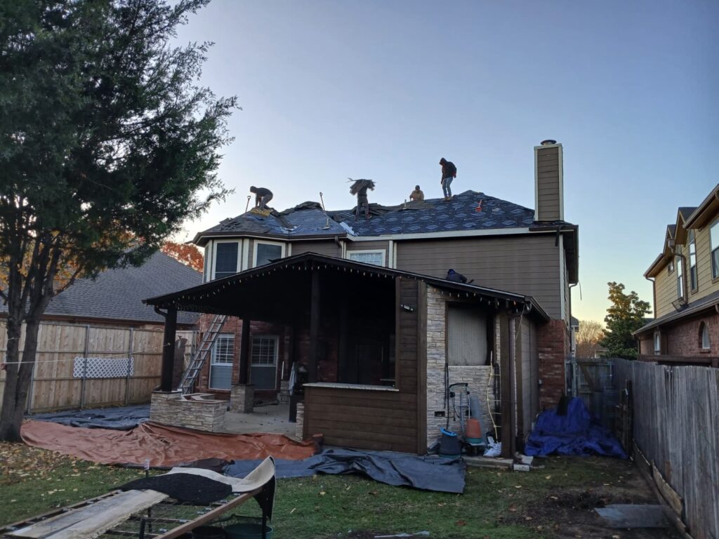 Several roofers are actively working on a residential roof, removing old shingles and installing new underlayment and asphalt shingles. Tarps are laid out on the ground around the house to catch debris.