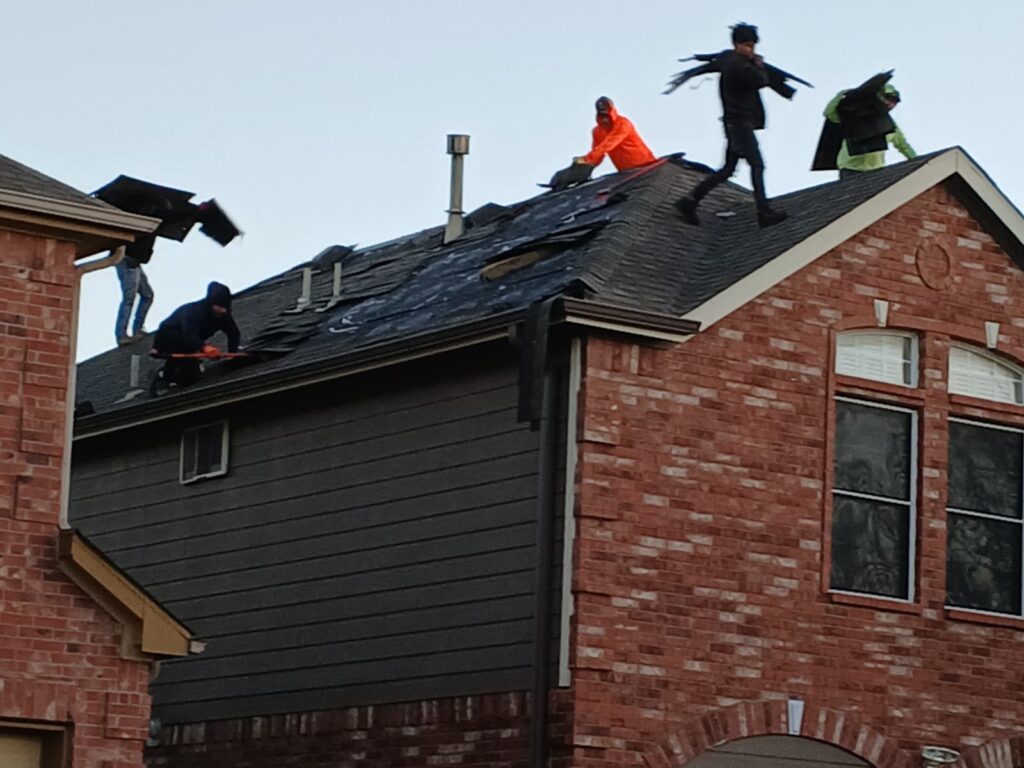 Several roofers are actively tearing off old asphalt shingles from a residential house roof. The roof is partially stripped, revealing underlayment and bare decking in some areas.