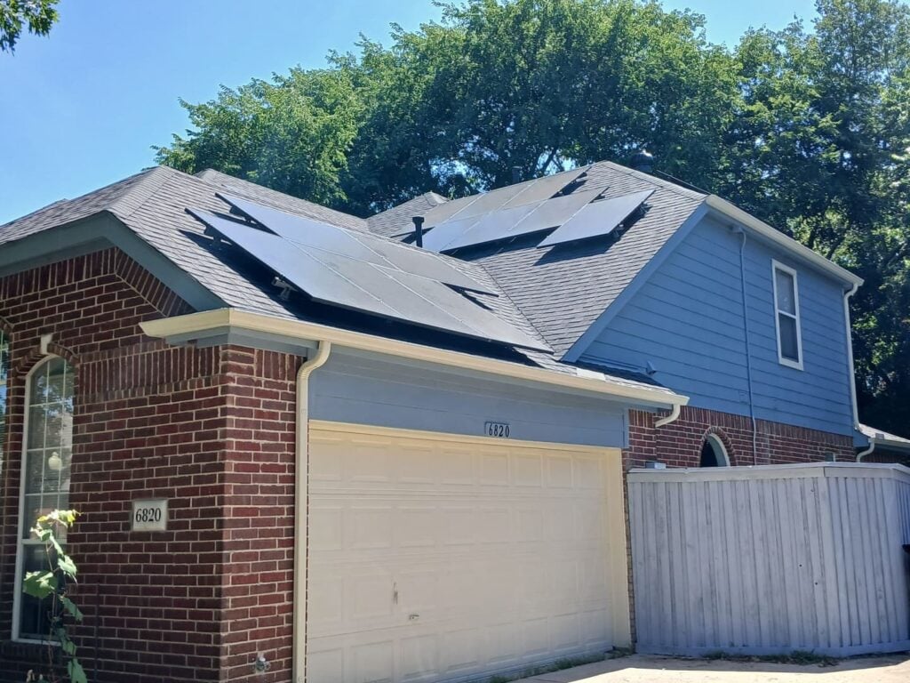 A residential house with a dark asphalt shingle roof, featuring several solar panels installed on multiple roof planes. The house has brick and blue siding, with a white garage door.