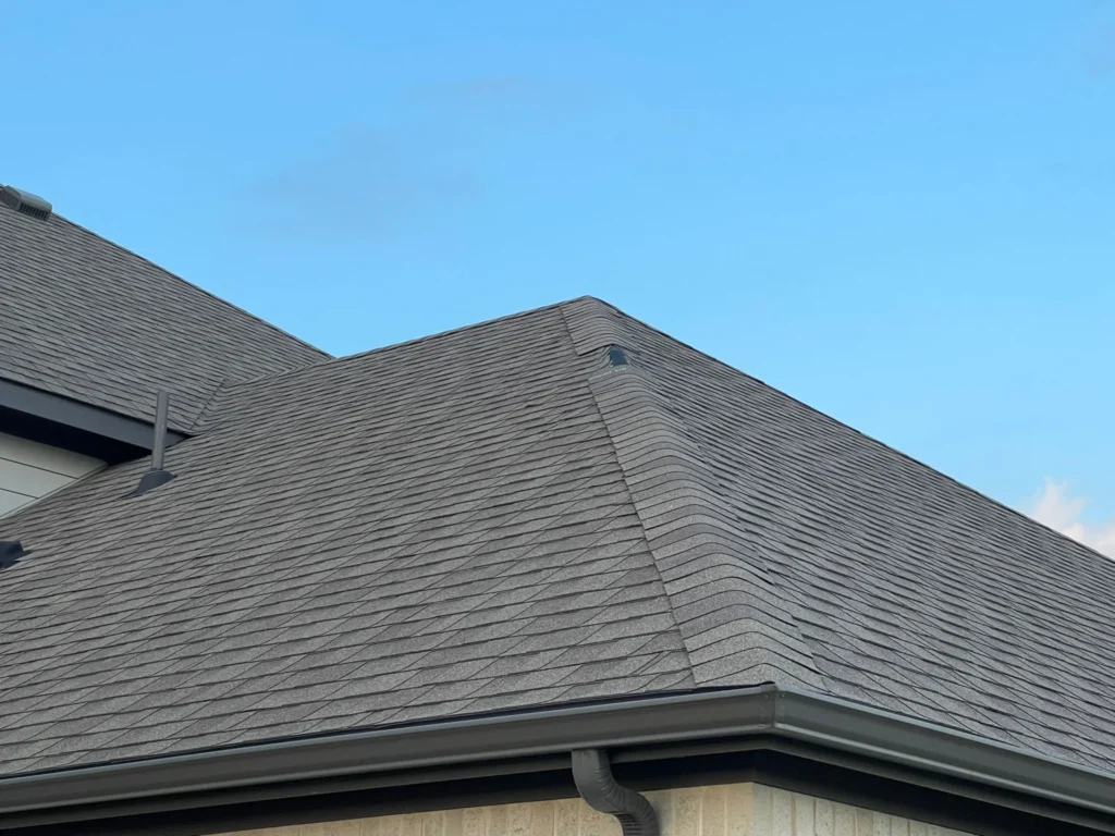 A section of a residential roof with dark gray asphalt shingles, dark gutters, and light brick siding, set against a clear blue sky.