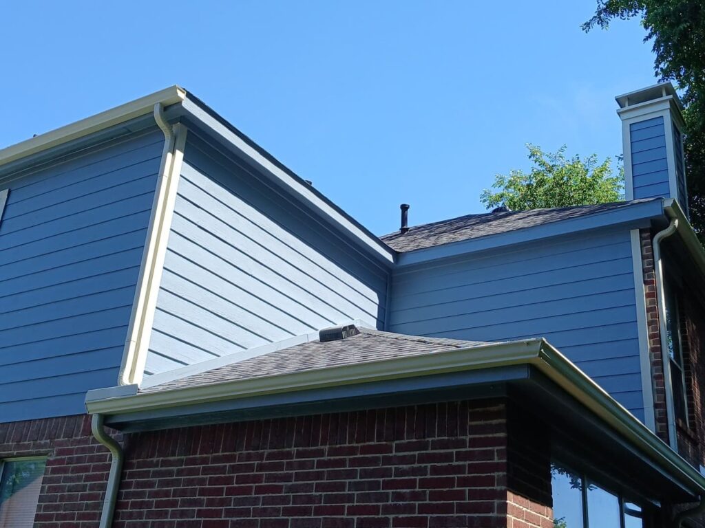 A residential house featuring blue siding, red brick, and an asphalt shingle roof, with gutters and a chimney visible against a clear blue sky.