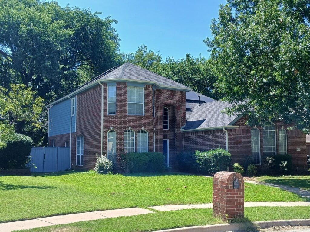 A two-story brick house with blue siding on one section, a dark gray asphalt shingle roof, and a well-maintained green lawn under a clear blue sky. Large trees frame the house, and a brick mailbox stands in the foreground.