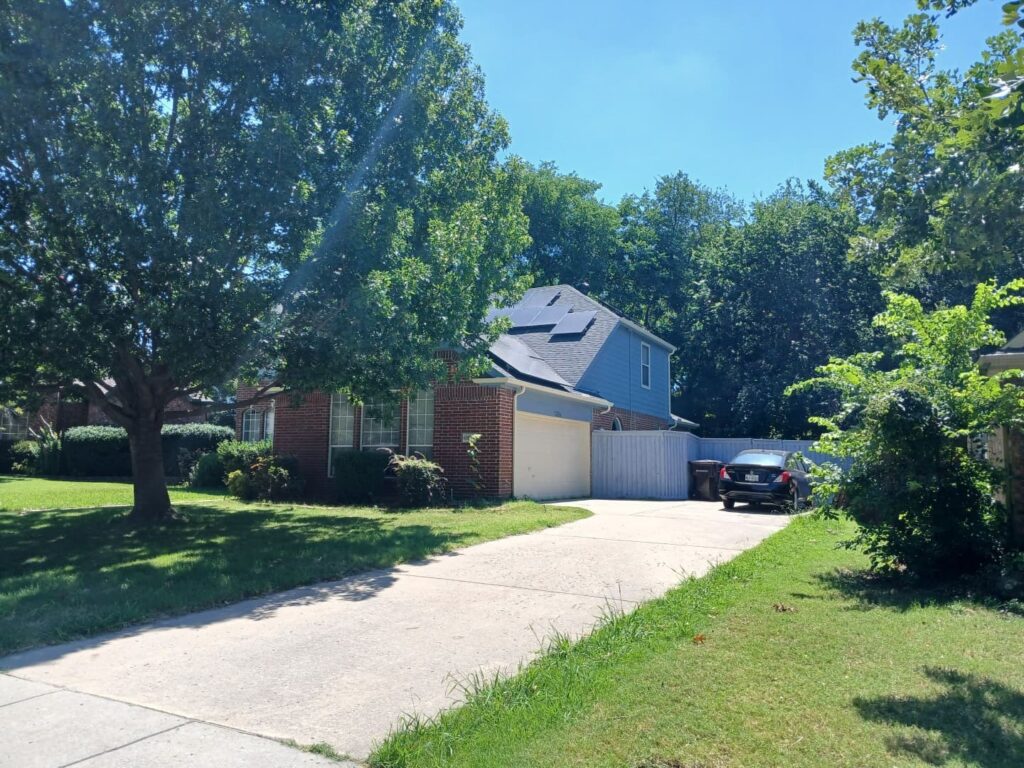 A residential house with a brick front and light blue siding on the upper story, featuring a dark asphalt shingle roof with several solar panels installed. A large tree partially obscures the front of the house and roof, with a concrete driveway leading to the garage.
