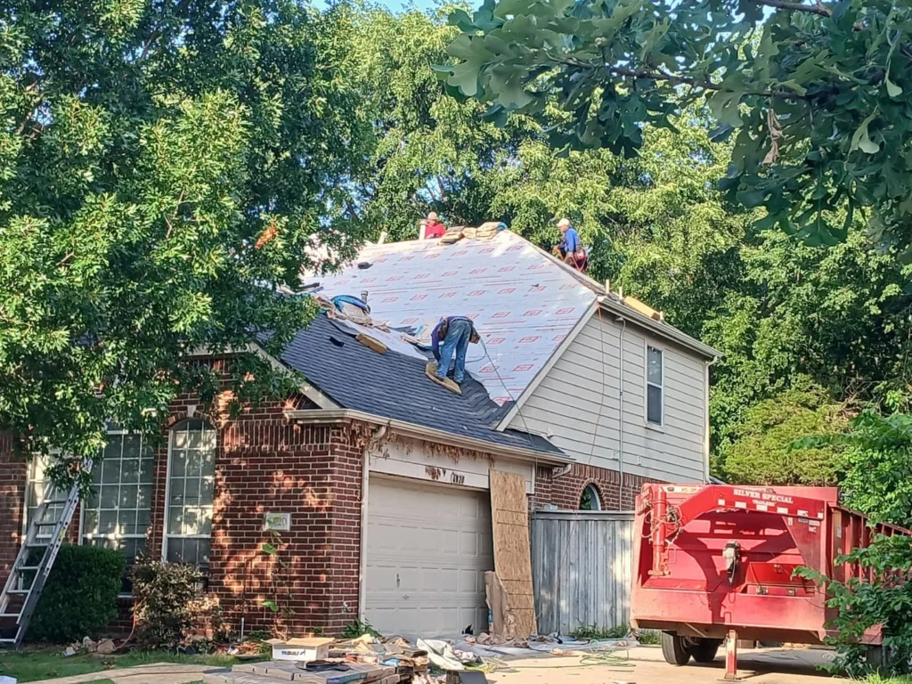 Several roofers are actively working on a residential roof, with some areas covered by underlayment and others still showing existing shingles. A large red dumpster is parked in the driveway, and a ladder leans against the house.
