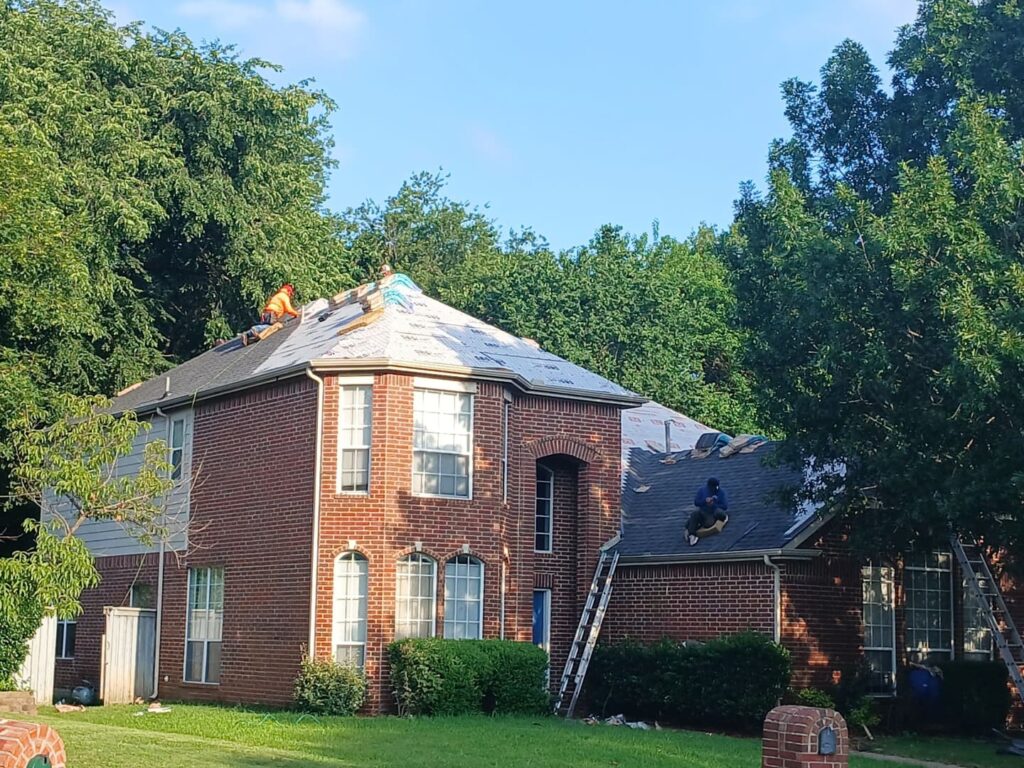 Two roofers are actively working on installing new asphalt shingles on a two-story brick residential house, with some sections of the roof still showing underlayment.