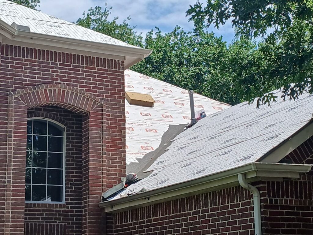 A close-up view of a brick house with a roof partially stripped and new underlayment installed. Green trees and a blue sky are visible in the background.