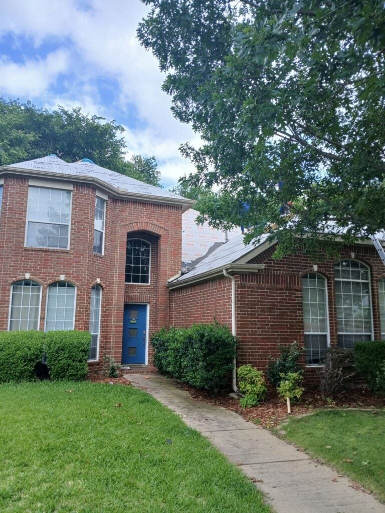 A two-story brick house with a blue front door, where the roof is currently being replaced. Underlayment is visible on the roof, and workers can be seen on the upper sections.