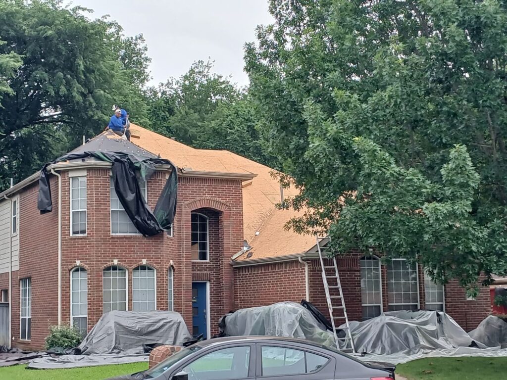 A brick residential house undergoing roof replacement, with a worker on the partially stripped roof and protective tarps covering the surrounding landscaping and ground.